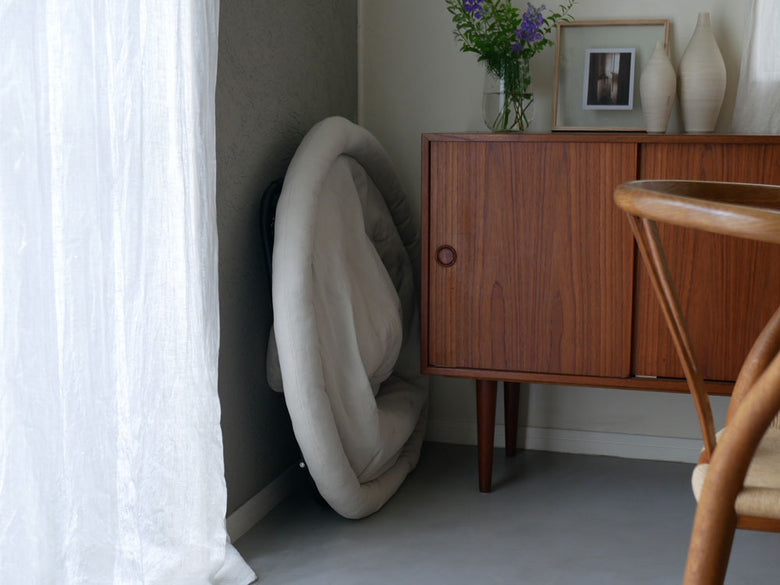 灰色の木目調床に置かれた【Comore Sun】2wayフォールディングチェアキャンバス。周囲にカーテンと観葉植物が配置された室内風景。